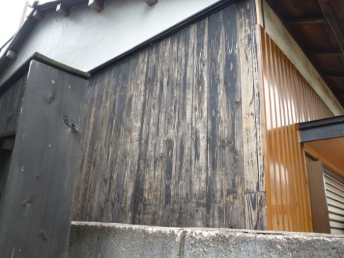 Weathered vertical wooden siding on a building, with a concrete base and orange metal panel to the right.