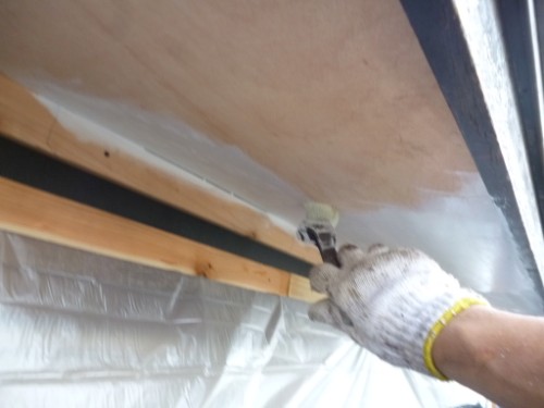Gloved hand applying white sealant with a small brush under a wooden beam, plastic sheeting visible in the background.