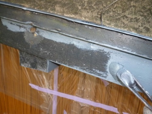 Close-up of a weathered metal beam with chipped paint, a cable resting on top, above a wooden paneled surface taped with pink painter’s tape.