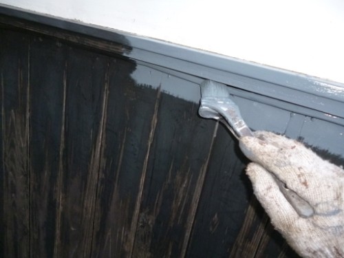 Close-up of a gloved hand brushing gray paint onto the trim along a dark wooden exterior siding.
