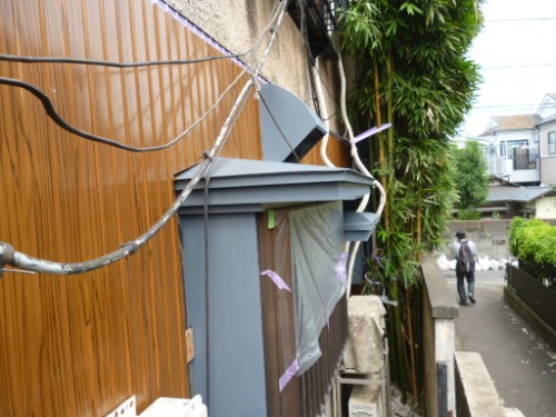 Residential alley with a wooden wall, exposed electrical wires, and a blue door; a man walks along the sidewalk in the distance.