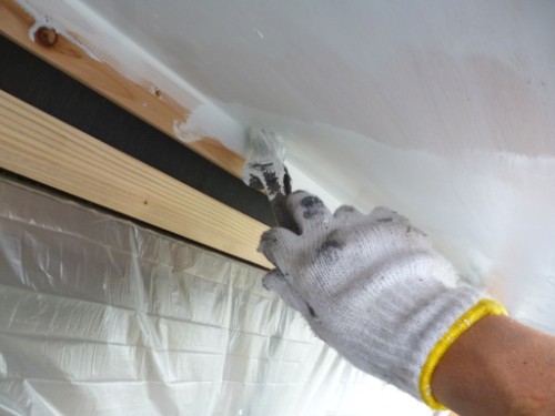 Close-up of a gloved hand smoothing white caulk along a seam between wall and ceiling, with plastic sheeting and a wooden strip visible in a renovation setup.