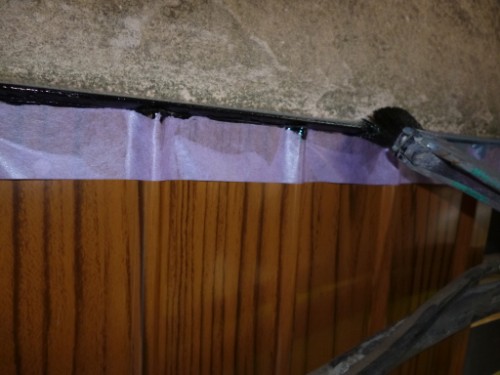 Close-up of a gap being sealed under a countertop with purple painter’s tape and a bead of black caulk.