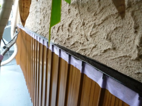 Close-up of a brown slatted railing with purple painter’s tape along the top edge, next to a textured beige wall, with a green leaf in the foreground.