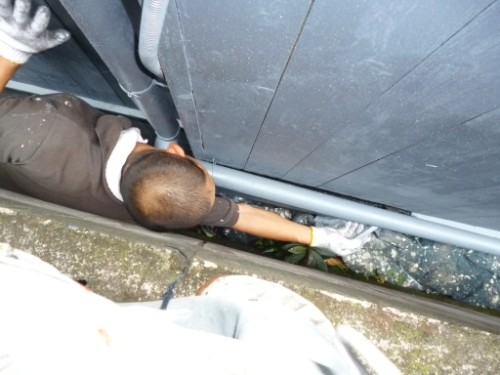 Worker wearing gloves reaching through a narrow gap between a wall and a pipe.
