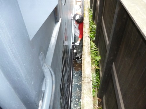 Worker in a red shirt and hard hat leaning over in a tight gap between a blue wall and a black fence, inspecting a surface near a large blue vehicle/structure.