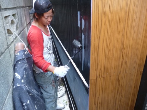 Person painting a black metal panel with a long roller, wearing a red-long-sleeve shirt and gloves.