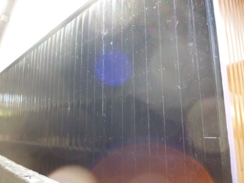 Dark glossy glass wall with vertical ridges and bright sun glare creating blue and orange lens flares in front of a wooden frame.