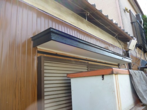 Back alley scene with a brown wooden wall, a metal roll-up door, and a white storage cabinet with an orange trim under an overhang.