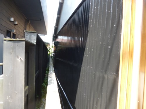 Narrow outdoor corridor between a wooden fence on the left and tall black metal fencing on the right, with a slim walkway in between.