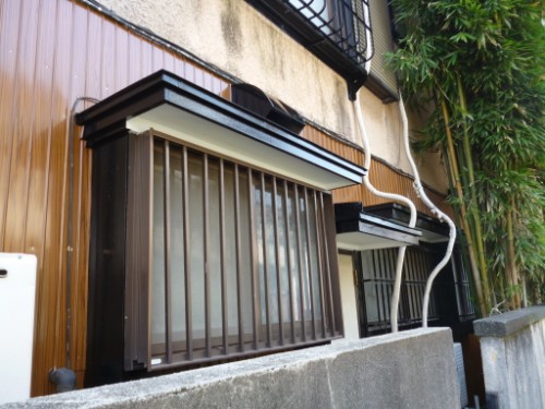 Exterior wall with metal security bars over windows, small brown window awnings, and exposed pipes along a beige building façade.