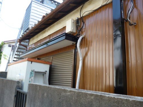 Exterior wall of a building with a white air conditioning unit, cables, and a brown wooden-siding facade near a concrete barrier.
