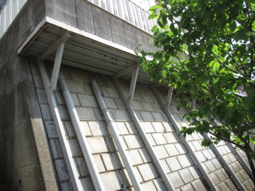 Concrete block wall with angled metal supports under a small overhang; green tree branches on the right.
