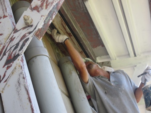 Construction worker wearing gloves applies sealant/caulk to a rusted metal beam around a doorway under an overhang.