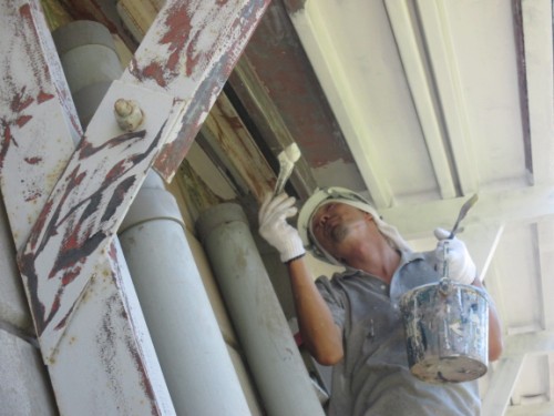 Worker wearing a cap and gloves, painting a rusted metal beam under an overhang with a paintbrush and can of paint nearby