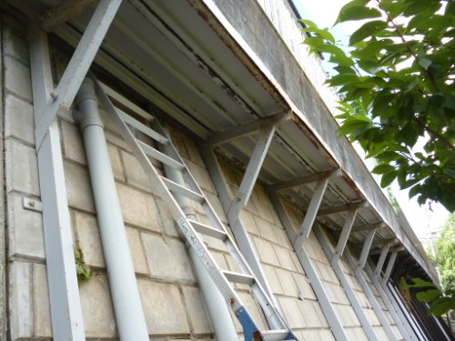 Ladder propped against a concrete wall beneath a metal walkway with supporting beams and pipes nearby