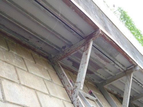 Rusty metal support brackets under a corrugated overhang attached to a brick wall.