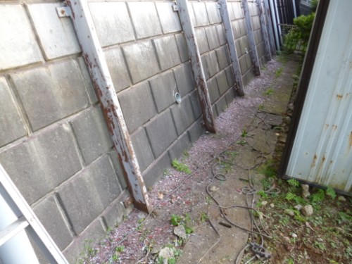 Narrow outdoor alley along a cinder block wall, with rusted vertical rails and a dirt path tangled with cables and weeds.