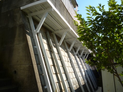 Underside of a raised walkway with white diagonal supports along a stone wall, with a tree on the right.