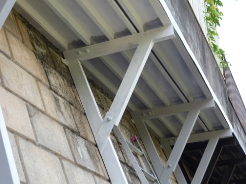Underside of a light gray corrugated metal canopy supported by diagonal braces attached to a concrete block wall.