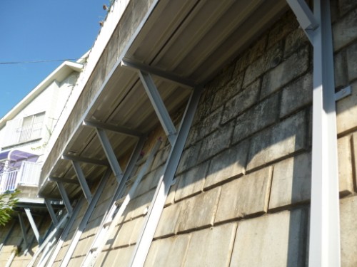 Low-angle view of a metal awning with grey support braces attached to a stone wall under a blue sky