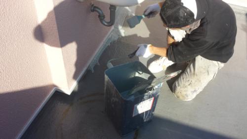 koushin_20140121tatiagari1 | 外壁塗装は神奈川県横浜市の塗装職人 Worker wearing gloves and a cap applies waterproof coating from a bucket onto a flat rooftop near a pink wall and vent pipe.