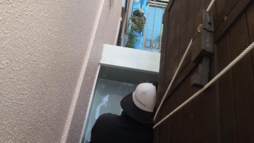 koushin_20140121uretan1 | 外壁塗装は神奈川県横浜市の塗装職人 Construction worker in a white hard hat stands in a narrow outdoor stairwell, looking up toward a person at the top near a gate/door.