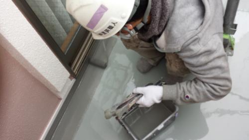 Worker in a helmet and gloves applying gray sealant along a balcony edge beside a sliding glass door.