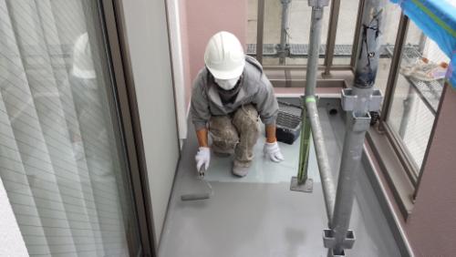 koushin_20140125top3 | 外壁塗装は神奈川県横浜市の塗装職人 Construction worker in a hard hat kneels on a balcony floor, applying a concrete patch with a trowel beside scaffolding and safety rails