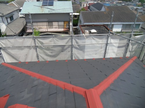 koushin_CIMG2586-1 | 外壁塗装は神奈川県横浜市の塗装職人 Rooftop under construction with dark gray shingles, red flashing at the ridge, and a white safety screen along the edge; houses visible in the background.
