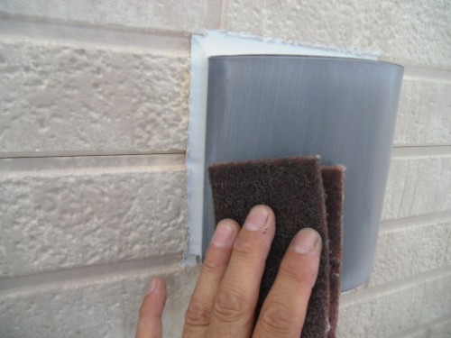 koushin_CIMG2607-1 | 外壁塗装は神奈川県横浜市の塗装職人 Hand holding a brown abrasive pad and wiping the caulked gap around a gray exterior vent cover on a beige textured wall