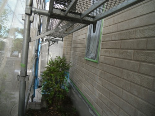 koushin_CIMG2612-1 | 外壁塗装は神奈川県横浜市の塗装職人 Scaffolding runs along the side of a beige brick house, with a taped window and a shrub beneath.