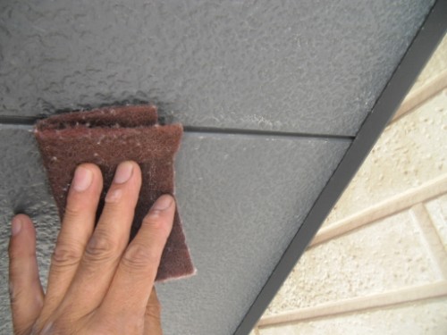 koushin_CIMG2615-1 | 外壁塗装は神奈川県横浜市の塗装職人 Hand holding brown sandpaper pad and rubbing a gray exterior panel, likely sanding the surface outdoors.