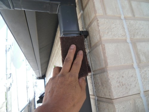 koushin_CIMG2616-1 | 外壁塗装は神奈川県横浜市の塗装職人 Close-up of a hand pressing a brown foam insulation block against a brick exterior near a gutter/downspout on a building.