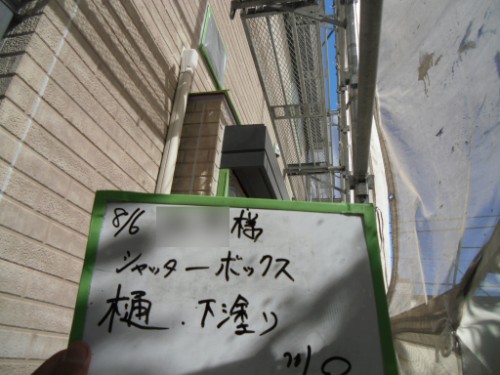 koushin_CIMG2624 | 外壁塗装は神奈川県横浜市の塗装職人 Construction site beside a brick wall; a hand holds a whiteboard with the date 8/6 and Japanese notes reading 'シャッターボックス' and '樋 下塗り' (shutter box and gutter undercoat). It shows scaffolding in the background.