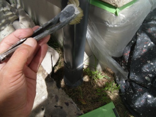 koushin_CIMG2626 | 外壁塗装は神奈川県横浜市の塗装職人 Person uses a brush to apply sealant or paint at the base of a metal post outdoors, near bags and debris on the ground