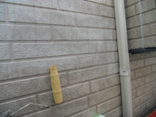 koushin_CIMG2635 | 外壁塗装は神奈川県横浜市の塗装職人 Yellow paint roller leaned against a beige brick exterior wall near a white downspout outside a house.
