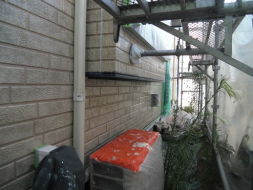 koushin_CIMG2636 | 外壁塗装は神奈川県横浜市の塗装職人 Narrow alley beside a brick wall with scaffolding, exposed pipes, and an orange-topped container; overgrown plants along the ground.