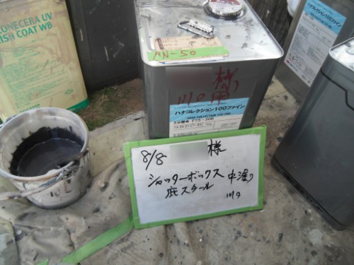 koushin_CIMG2638 | 外壁塗装は神奈川県横浜市の塗装職人 Outdoor scene with several metal paint cans and a bucket containing dark liquid; a whiteboard with handwritten Japanese text leans against one can, suggesting a mixing or labeling area for coating materials.