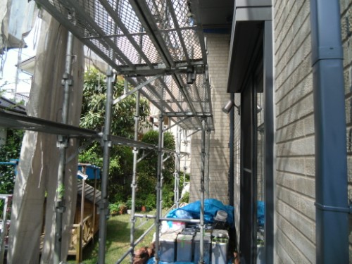 koushin_CIMG2641 | 外壁塗装は神奈川県横浜市の塗装職人 Scaffolding erected along the side of a brick building with metal planks and blue tarps covering materials under a sunny sky.