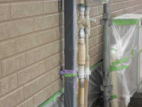 koushin_CIMG2646 | 外壁塗装は神奈川県横浜市の塗装職人 Exterior wall showing beige utility pipes and valves mounted to a vertical conduit, wrapped in plastic sheeting and green tape during installation.