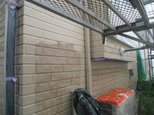koushin_CIMG2647 | 外壁塗装は神奈川県横浜市の塗装職人 Exterior brick wall under renovation with scaffolding and protective plastic coverings; a downspout runs along the wall and a bag rests on the ground near an orange-topped workbench.