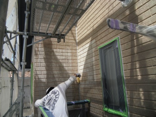 koushin_CIMG2654 | 外壁塗装は神奈川県横浜市の塗装職人 Worker on scaffolding painting a brick exterior wall with a roller; window is masked with green painter’s tape.