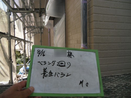 koushin_CIMG2663 | 外壁塗装は神奈川県横浜市の塗装職人 A person holding a small whiteboard with handwritten Japanese text, outdoors beside a brick wall with scaffolding in the background.