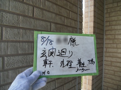 koushin_CIMG2668 | 外壁塗装は神奈川県横浜市の塗装職人 Gloved hand holding a small whiteboard with handwritten Japanese text, mounted on a brick exterior wall with a green border.