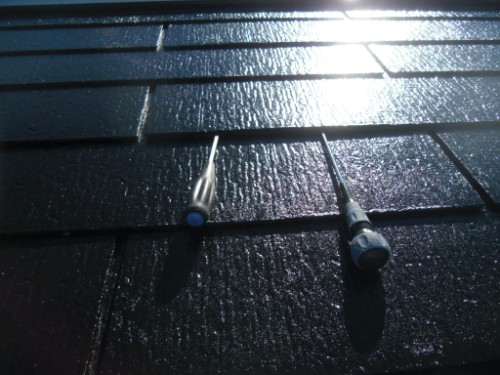 koushin_CIMG2681 | 外壁塗装は神奈川県横浜市の塗装職人 Two metal roof anchors with blue caps attached to dark shingles, with sun glare above.