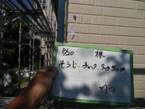 koushin_CIMG2685 | 外壁塗装は神奈川県横浜市の塗装職人 Person holding a small whiteboard with handwritten Japanese text outdoors beside a brick wall and metal stairs/railings.