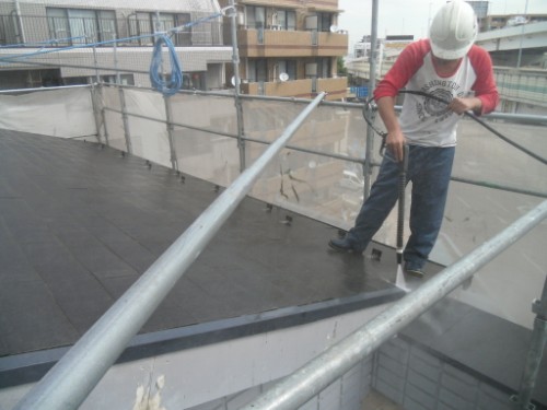 koushin_CIMG2873-1 | 外壁塗装は神奈川県横浜市の塗装職人 Construction worker wearing a helmet on a rooftop, using a tool near metal safety rails with urban buildings in the background.