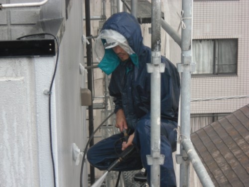koushin_CIMG2883-1 | 外壁塗装は神奈川県横浜市の塗装職人 Maintenance worker in a blue rain suit on scaffolding, using a tool on the exterior of a building.