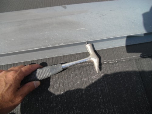 koushin_CIMG2923-1 | 外壁塗装は神奈川県横浜市の塗装職人 Hand holding a hammer-like tool and tapping a metal roof seam on a dark roof surface.